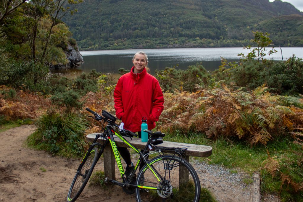 Woman wearing a red jacket smiles at the camera while standing next to a bike with a water bottle on the bench behind her is a lake and mountain while biking in Killarney National Park.