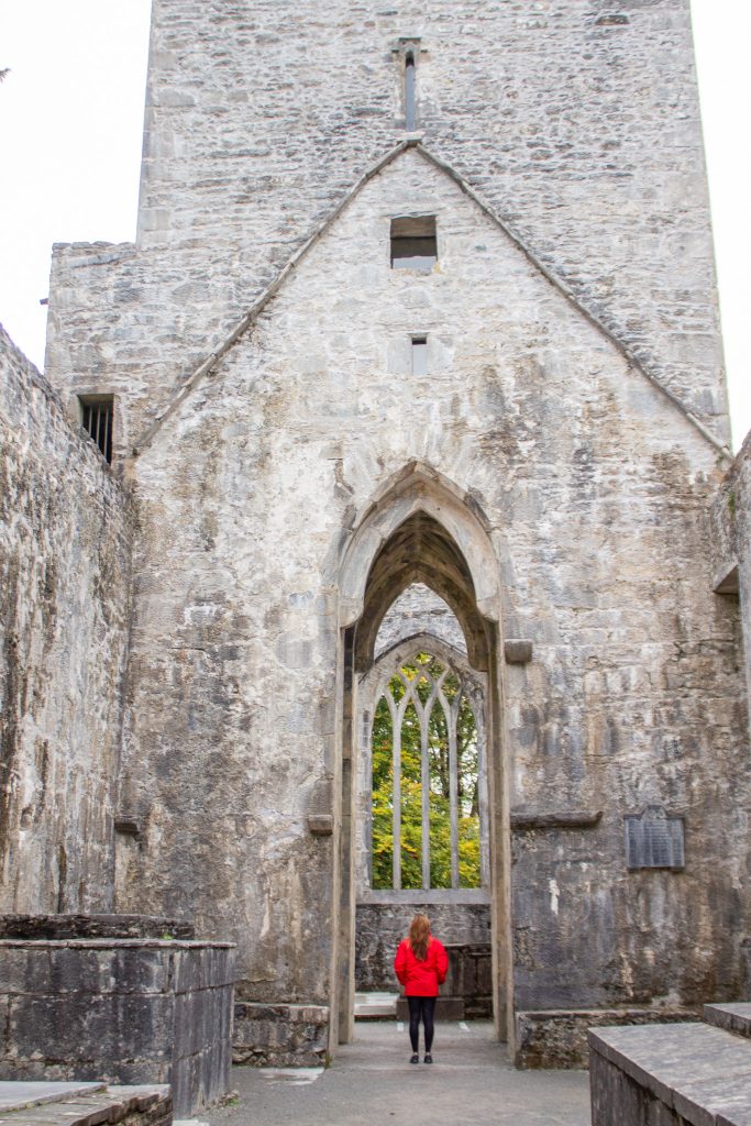 A woman wearing a red jacket stands beneath an stone arch entry way for an old, ruined abbey.