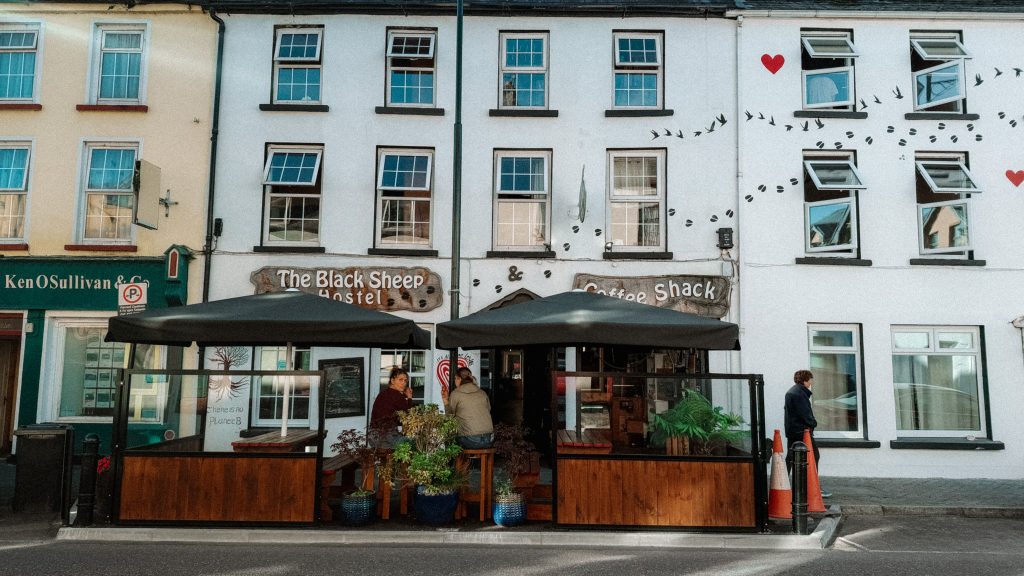 White building that says Black Sheep Hostel with two umbrellas in front with people sitting at stools underneath.