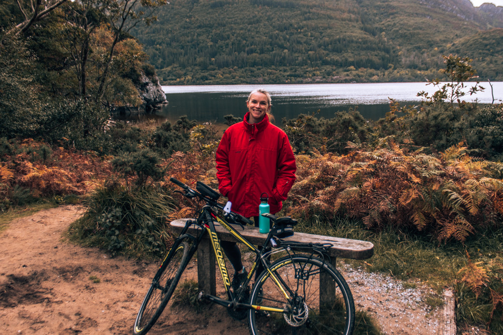 Katie standing beside a lake with a bicycle and water bottle while cycling through Killarney National Park.