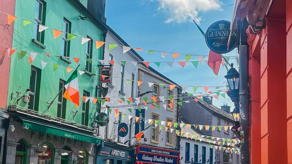 Colorful shops in Galway decorated with orange white and green Irish flags on a sunny day.