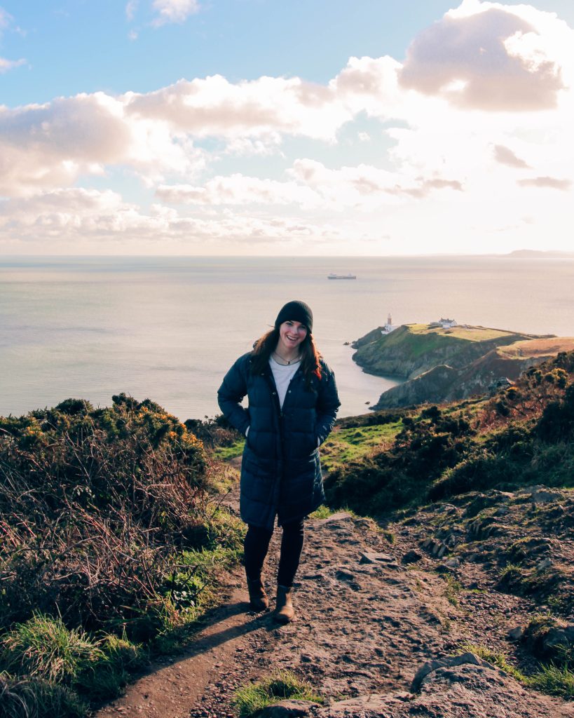 Katie walking along the Howth cliff path with a lighthouse visible in the distance and the coastline beyond.