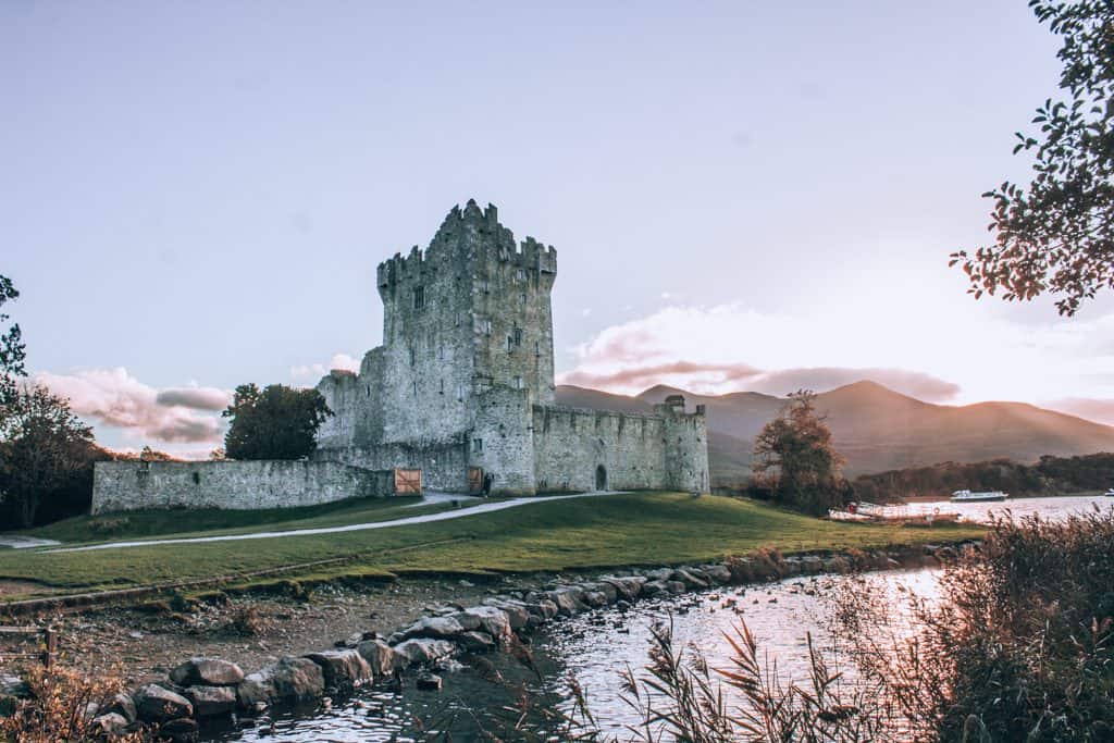 A white stone castle sits on green grass next to a lake as the sun sets in Killarney National Park.