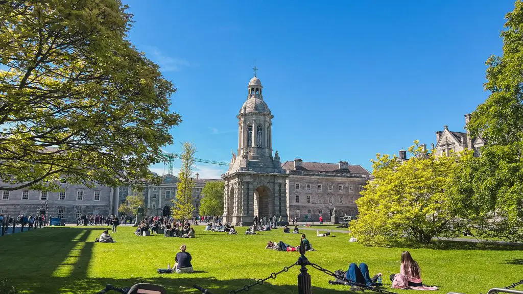 Trinity College Dublin courtyard on a sunny day with people sitting on the green lawn around the historic stone campanile. The landmark is part of the suggested two night Dublin stay.