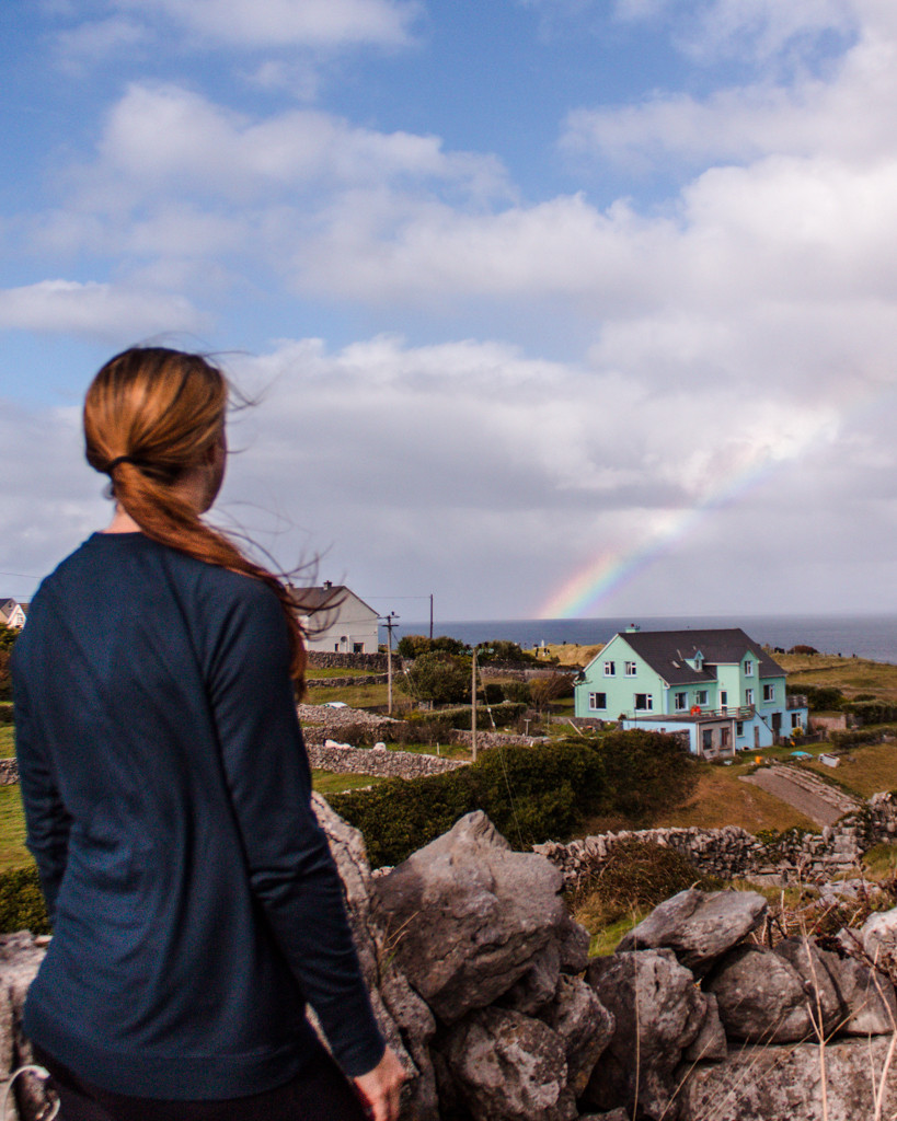 Katie overlooking the Aran Islands with green hills, rocky coastline, and a rainbow in the distance.