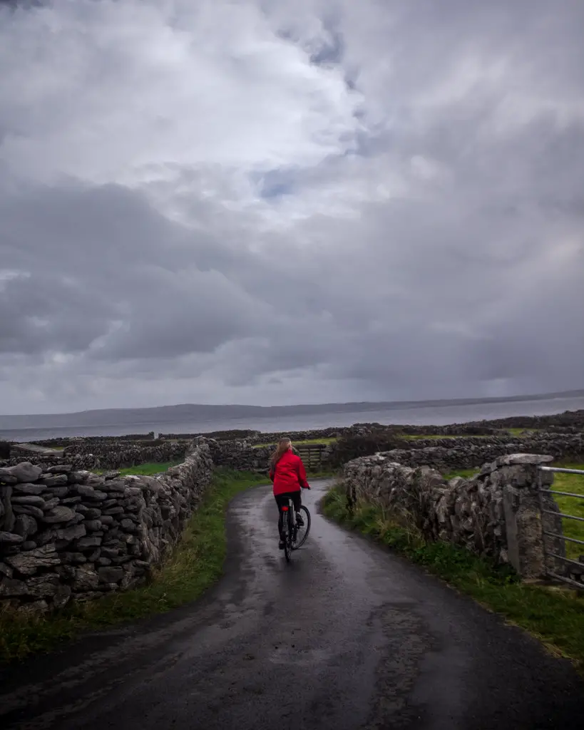 Katie riding a bike down a path between stone walls on a stormy day on Inisheer Island in Ireland.