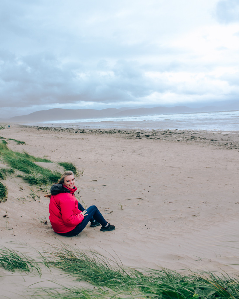 Katie wearing a red jacket and sitting on a white sand beach in Ireland on a stormy day.
