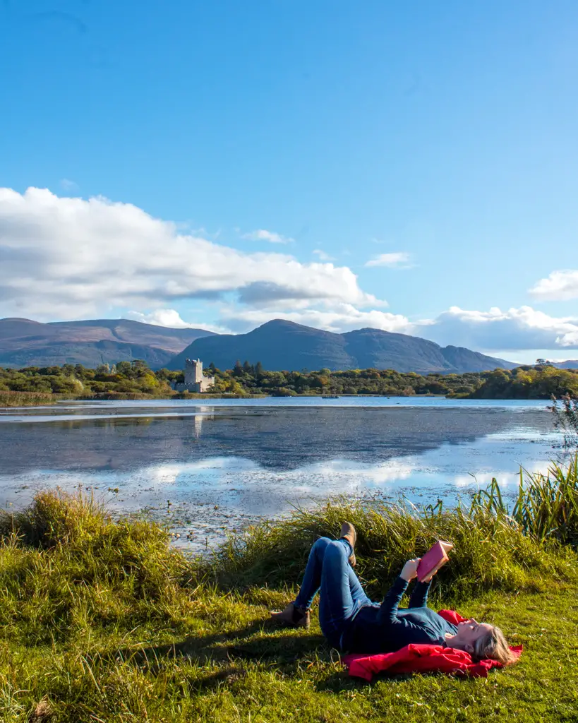 Katie laying on grass reading a book with Ross Castle in the distance in Killarney National Park in Ireland.