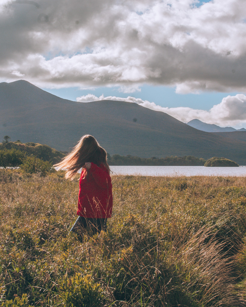 Katie standing in tall grass in front of the lakes of Killarney as the sun sets.