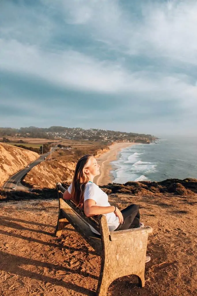 Woman sitting on a bench overlooking the Pacific Ocean and Half Moon Bay in the distance at sunset. 