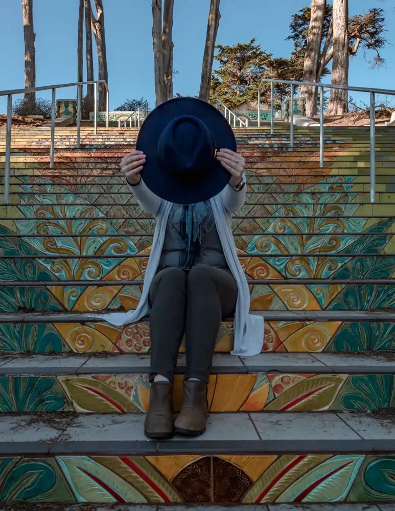 woman sitting on tiled steps holding hat in front of face