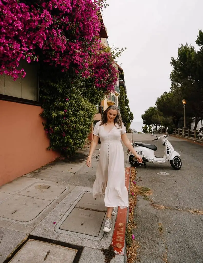 Walking down a street on Telegraph Hill with a vespa in the background wearing a white dress.