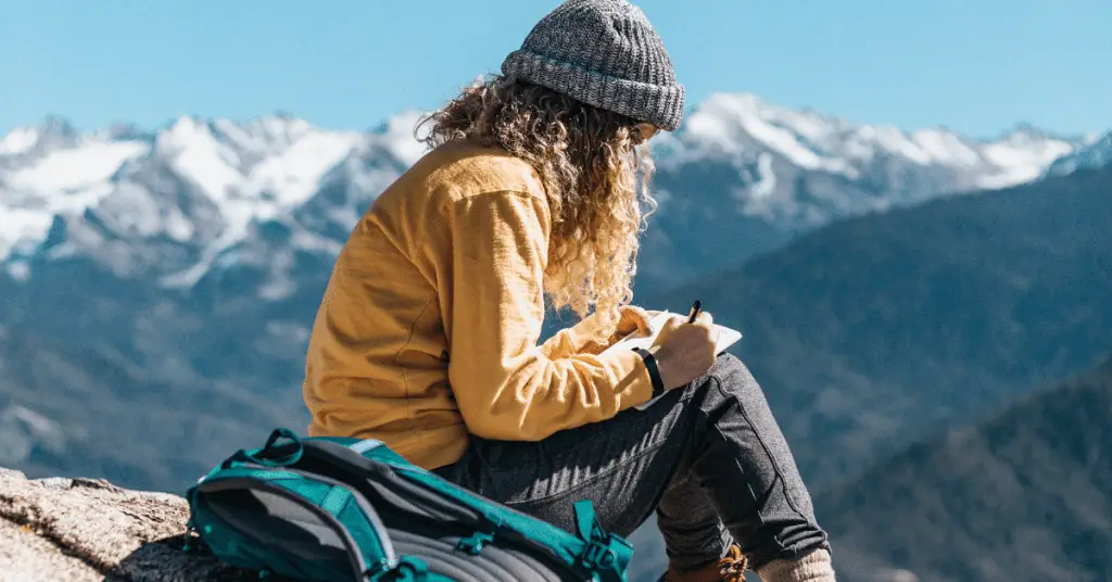 Planning a solo trip requires a solid bucket list. Image of a woman sitting on a mountain top journaling.