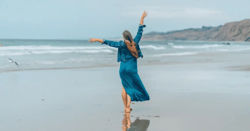 Travel with confidence by purchasing travel insurance before your flight. Image of a woman walking on a beach with her hands in the air. 