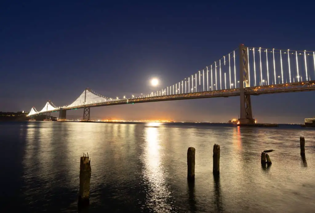 Bay Bridge lit up at night with the full moon reflected on the water below.