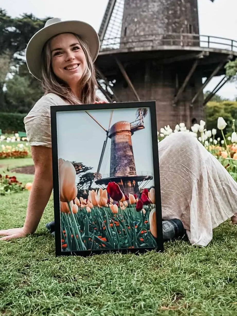 Wearing a white dress and hat sitting on the grass in front of the Golden Gate Park windmill holding a photo of the windmill.