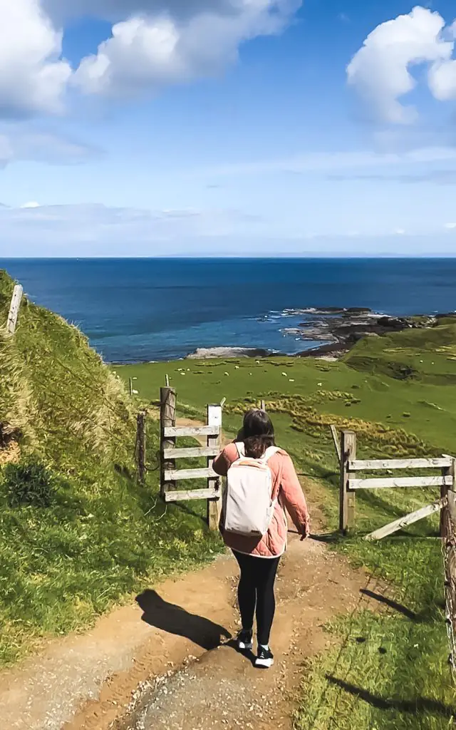Me walking down a dirt bath surrounded by green fields towards the blue sea wearing Hokas, leggings, a pink jacket and my backpack.