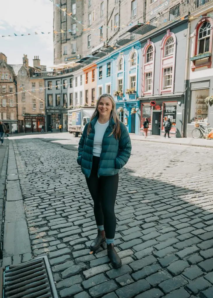 Me standing on Victoria Street in Edinburgh Scotland wearing a blue puffy jacket, white tank top, black leggings, and blundstone boots.
