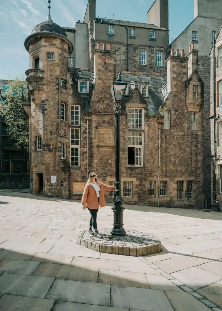 Me holding onto a lampost in front of the writer's museum in Edinburgh Scotland wearing a pink coat and black leggings. 