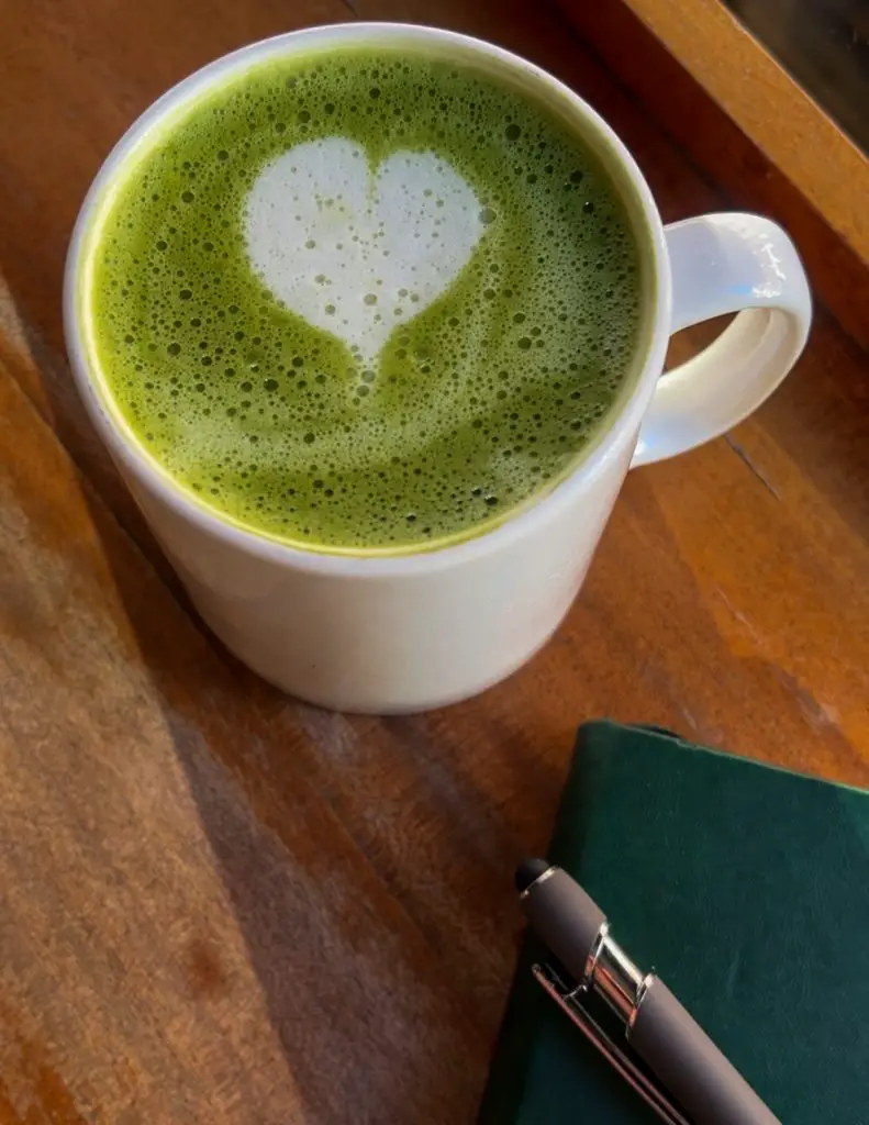 Green matcha latte in a white mug on a wood table with a green journal and pen next to it. 
