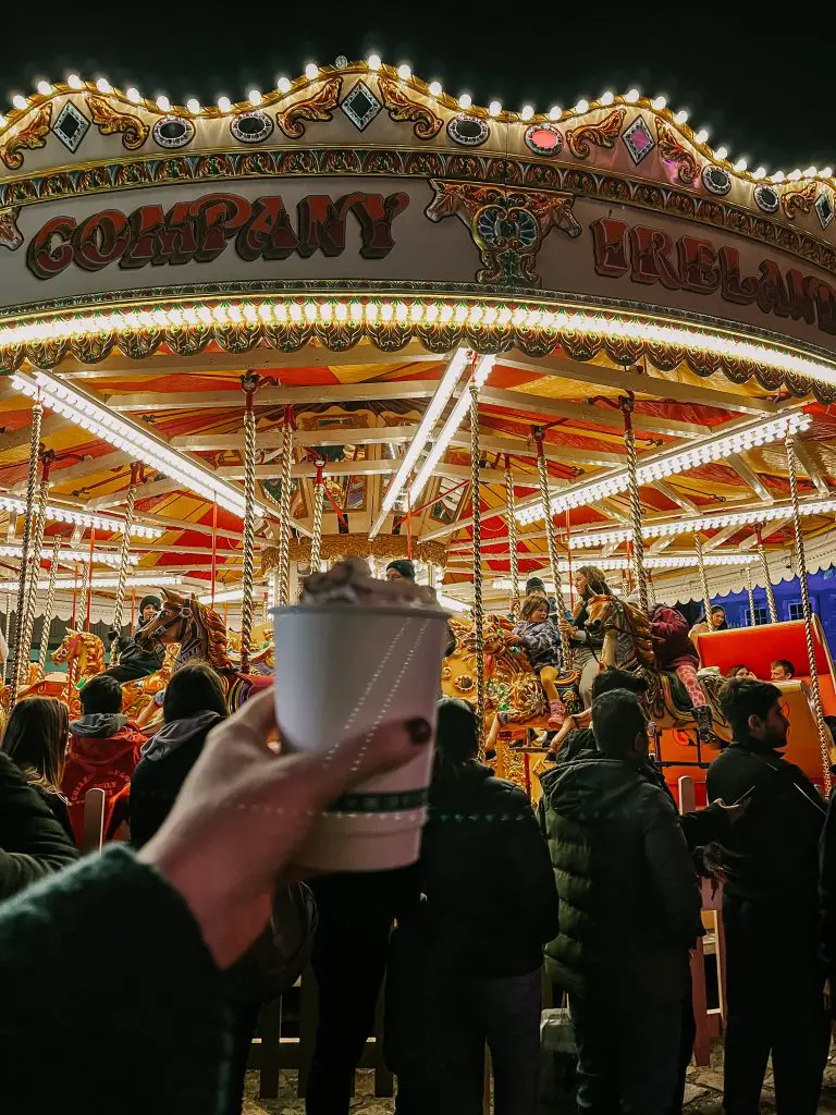 Holding a hot chocolate up in front of the carousel at the Dublin Castle Christmas market. 
