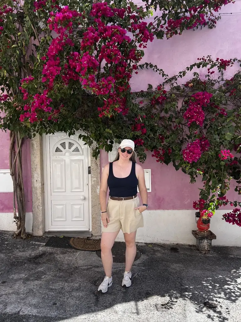 Wearing trouser shorts and a blue tank top in front of a pink building with pink flowers in Lisbon Portugal.