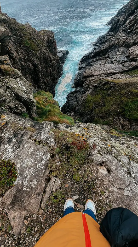 A hiker in an orange jacket stands near the edge of rocky cliffs overlooking turquoise waves crashing through a narrow coastal inlet in Donegal. The steep terrain highlights why careful footing is important when hiking Ireland's coastline.