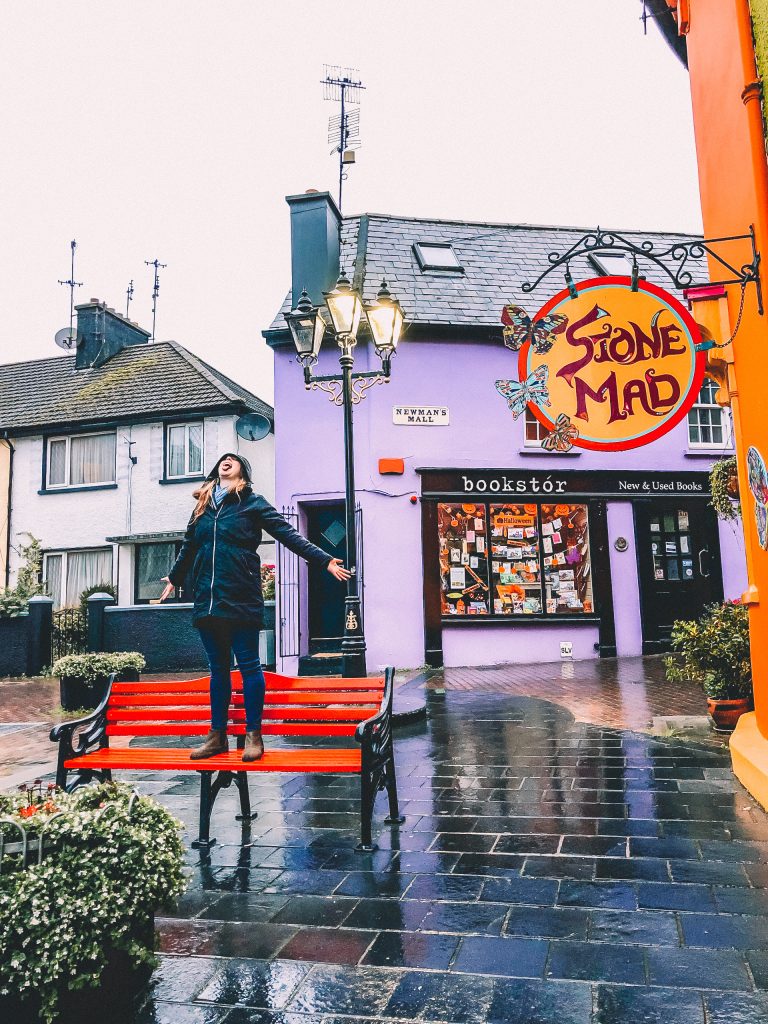 Katie standing on a bench in the colorful town of Kinsale in the rain.