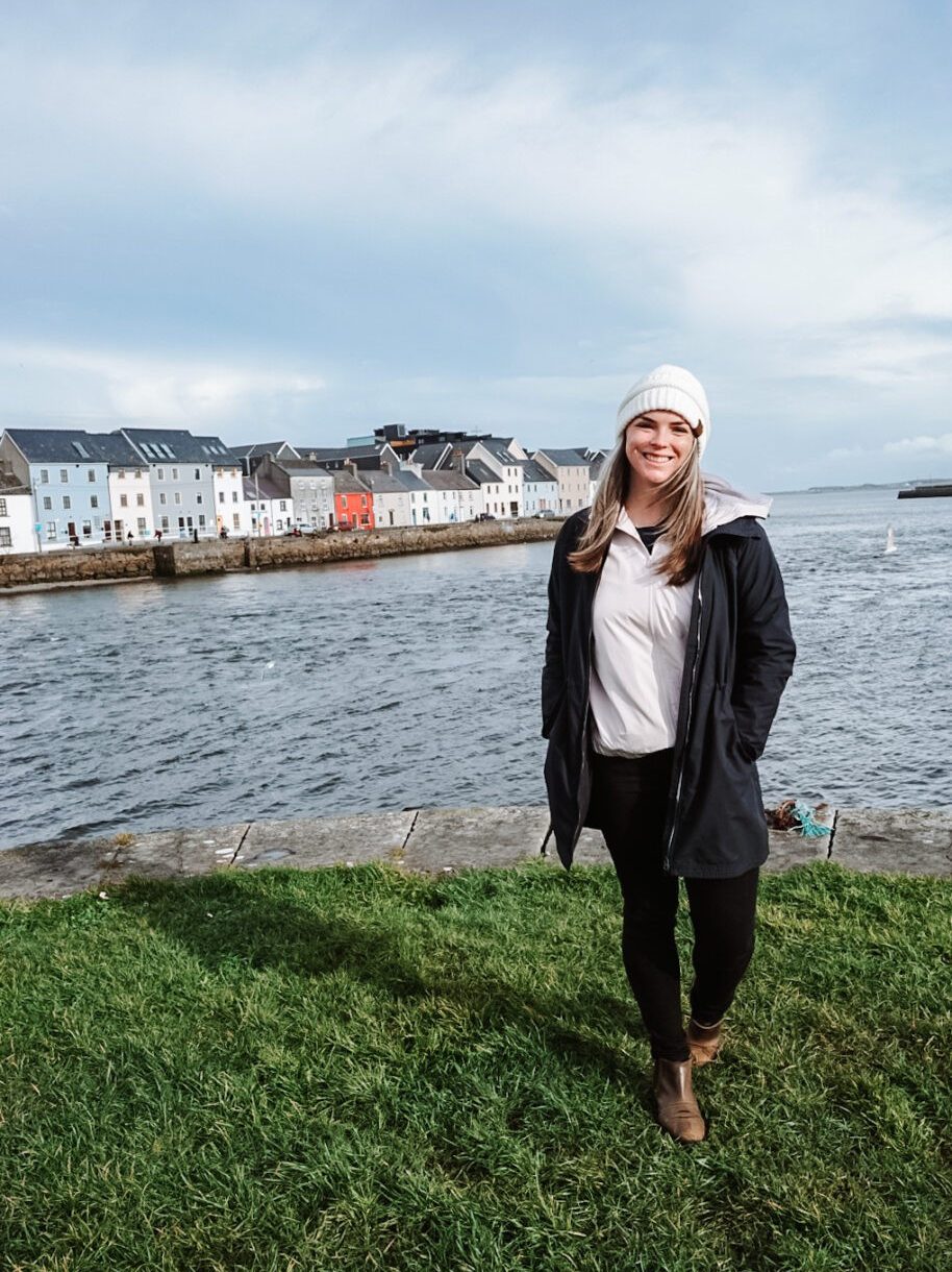 Katie standing on a grassy patch in front of Galway Bay and the colorful houses.