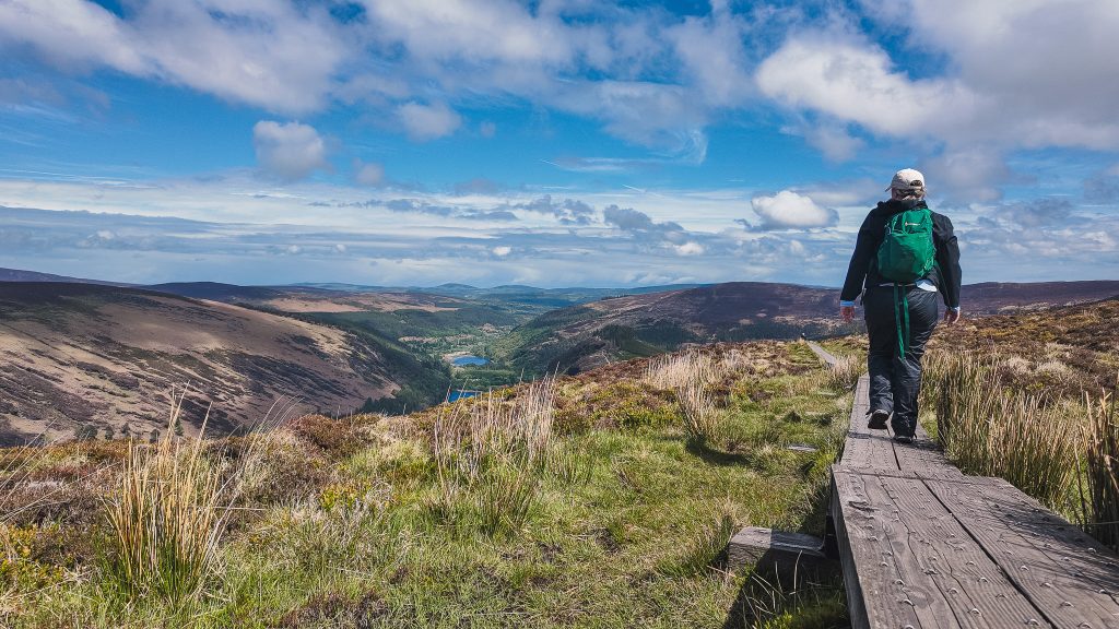 Katie wearing all black rain gear and a green backpack walks on an elevated boardwalk path surrounded by boggy ground and a lake off in the distance in Glendalough, Ireland.