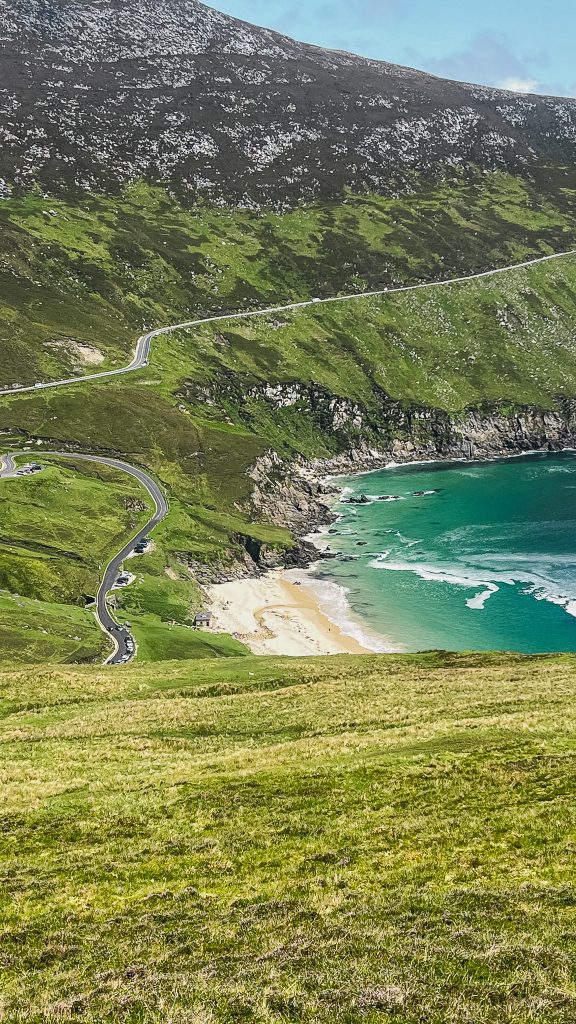 Turquoise waters of Keem Bay surrounded by steep green mountains and rocky cliffs, with a winding road leading down to the sandy beach. This scenic view highlights one of the most beautiful places to visit on Achill Island, Ireland.