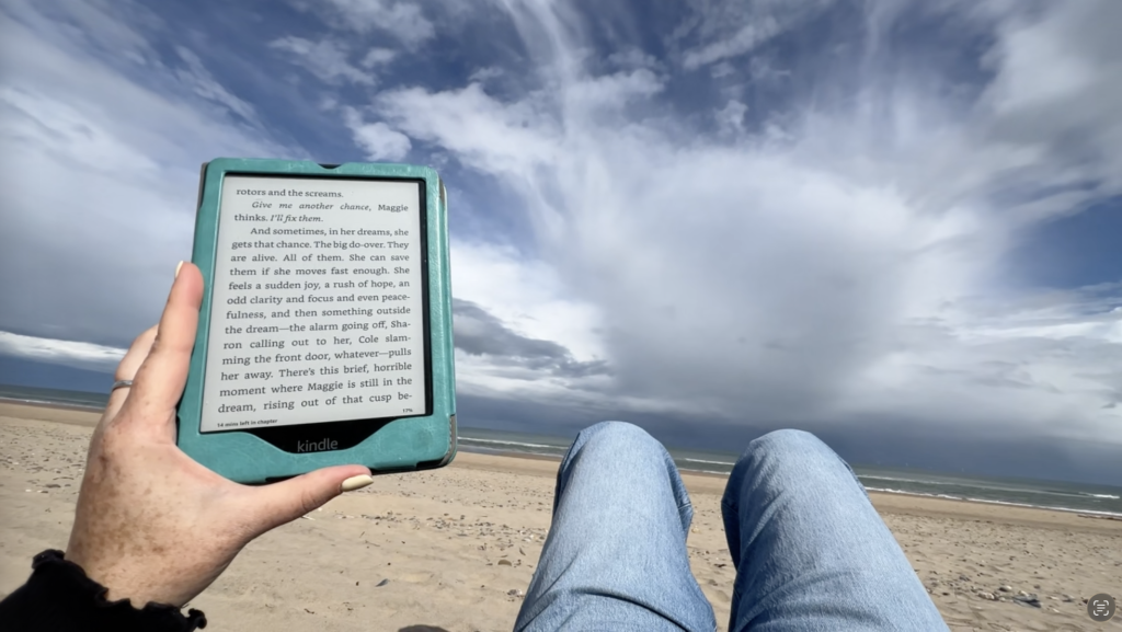 A hand holds a kindle with writing on it while two legs wearing jeans poke up with a cloudy blue sky over the beach in Ireland.