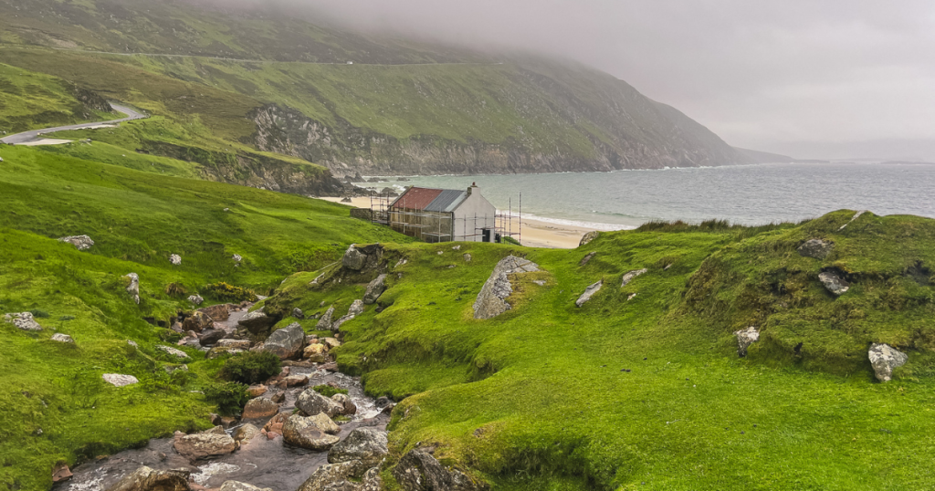 Small creek surrounded by green hills lead to a cottage overlooking a beach called Keem Bay on a stormy day.