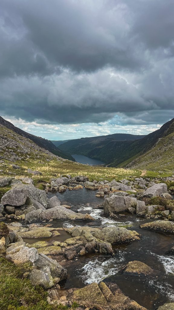River running through rocks leading to a lake surrounded by mountains and a stormy sky on the Spinc Trail in Glendalough, Ireland.