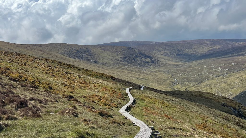 Boardwalk trail leading into a mountain landscape under stormy skies in Glendalough, Ireland.