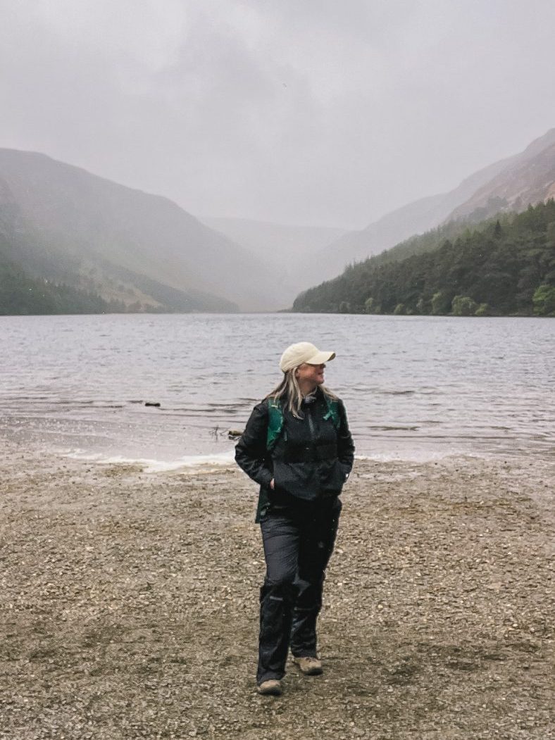 Katie wearing all black standing on a sandy beach in front of a lake with the Wicklow mountains in the distance.