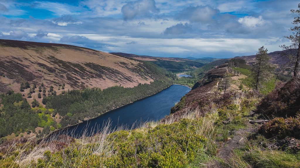 View of a long blue lake beneath mountains on a sunny day in Glendalough, Ireland.