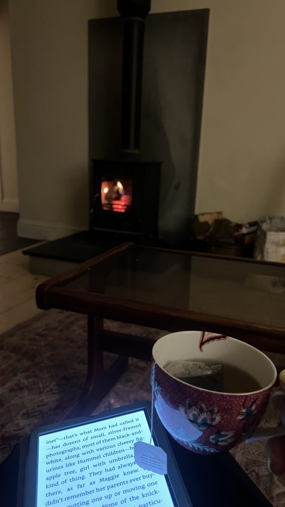 Kindle lit up with a cup of tea next to it and a stove fireplace lit up in the background.