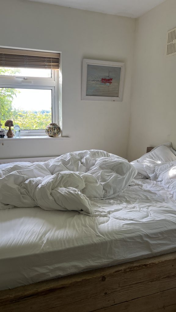 Messy bed with white bed sheets below a window with green trees outside.