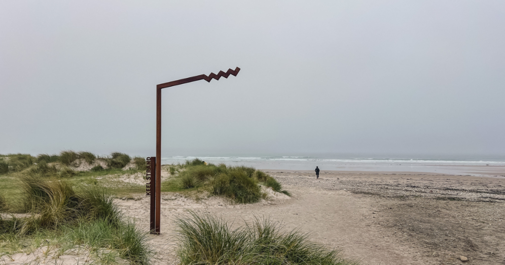 A wide sandy beach on Achill Island with grassy dunes in the foreground, a lone person walking toward the Atlantic, and a rusty metal sculpture standing near the shore under a cloudy sky.