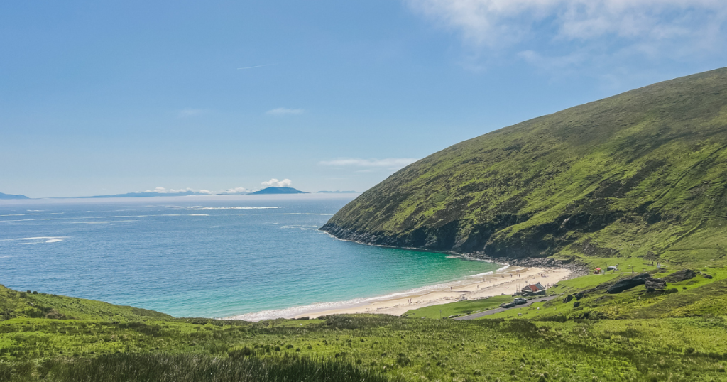 Green cliffs lead to a white sand beach and turquoise water with a small cottage on Achill Island.