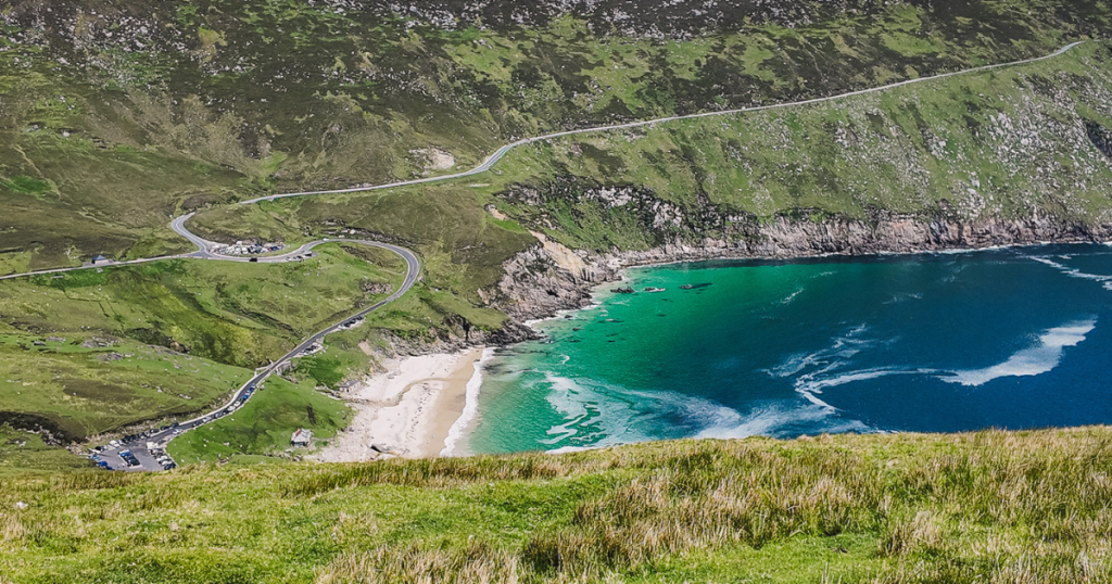 Turquoise waters of Keem Bay surrounded by steep green mountains and rocky cliffs, with a winding road leading down to the sandy beach. This scenic view highlights one of the most beautiful places to visit on Achill Island, Ireland.