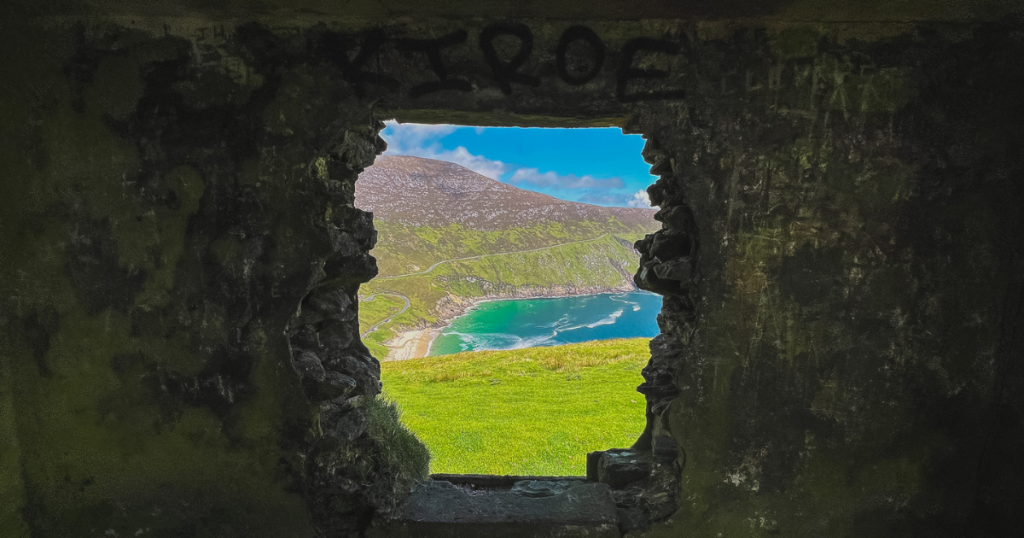 Keem Bay viewed through a rough opening in the stone walls, with bright green grass, turquoise water, a winding coastal road, and steep mountains beyond.