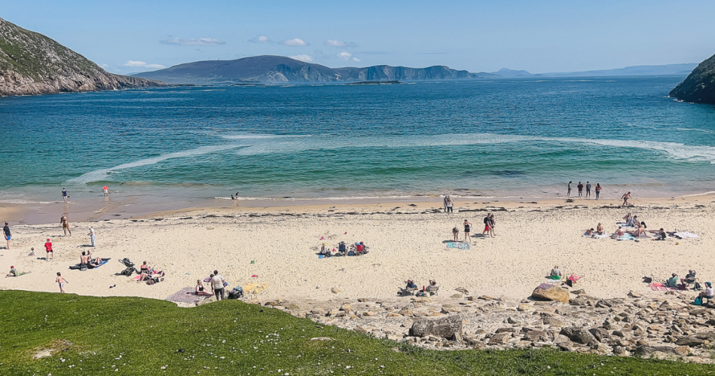 Multiple groups of people sitting on a the white sand beach of Keem Beach with turquoise water and mountains in the distance on a sunny day.