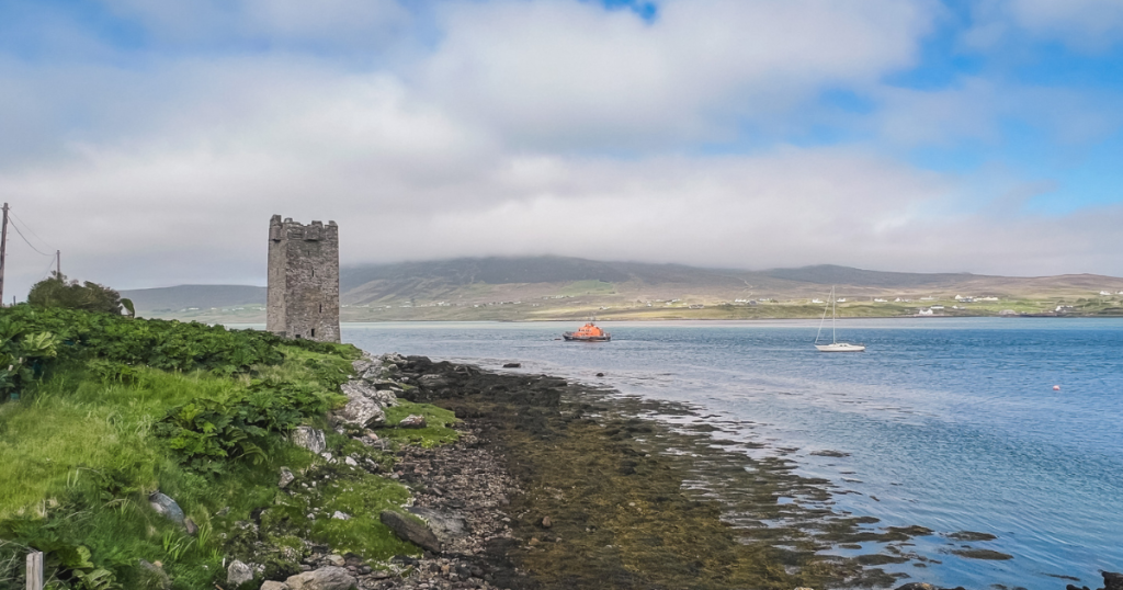Tower castle stands on the shores of Achill Sound with mountains in the distance.