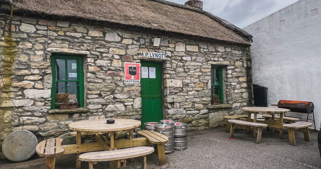 Small stone pub with picnic tables outside, a green door, and kegs in the front.