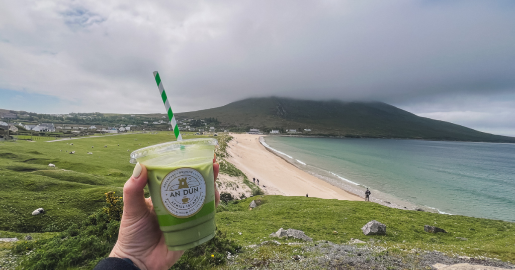 Hand holding a green iced matcha latte in front of Dugort beach on a foggy day on Achill Island.