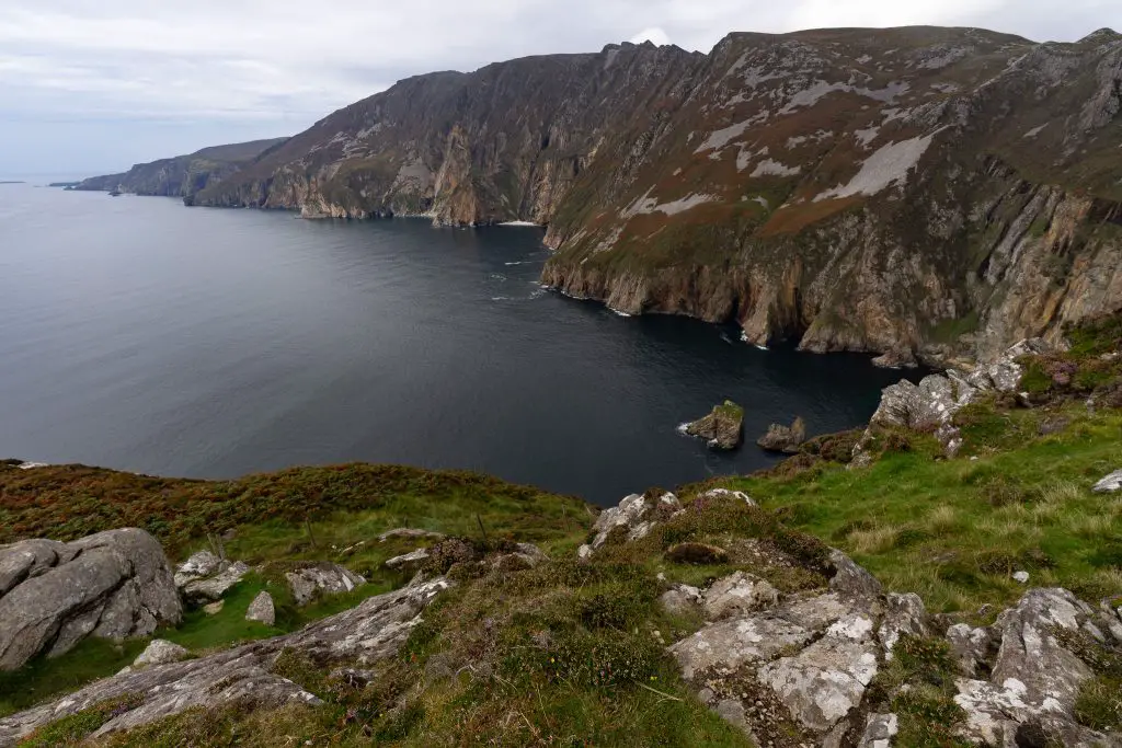 Dramatic green and brown cliffs rise above dark blue water at Slieve League in County Donegal. Rocky terrain fills the foreground beneath an overcast sky.
