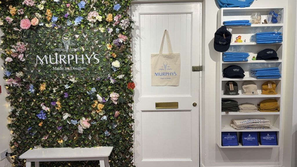 On the left side of the wall is greenery with a sign that says Murphys, next to it is a white door, and next to that are shelves filled with shirts and hats for sale.