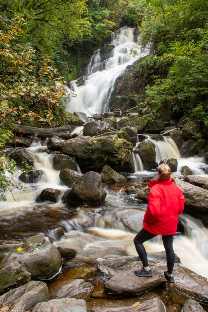 Woman wearing a red jacket and black leggings stands on rocks looking at a waterfall that flows over rocks between lush greenery.
